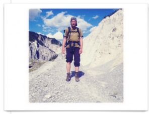 Wanderer mit Rucksack (Irgendlinck, also ich) posiert vor fast weißem Geröll und blauem Himmel. Das Bild ist quadratisch auf Postkarte, weiß