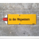 Ein gelbes Wanderwegschild, auf dem geschrieben steht 'zu den Wegweisern', zeigt nach links.